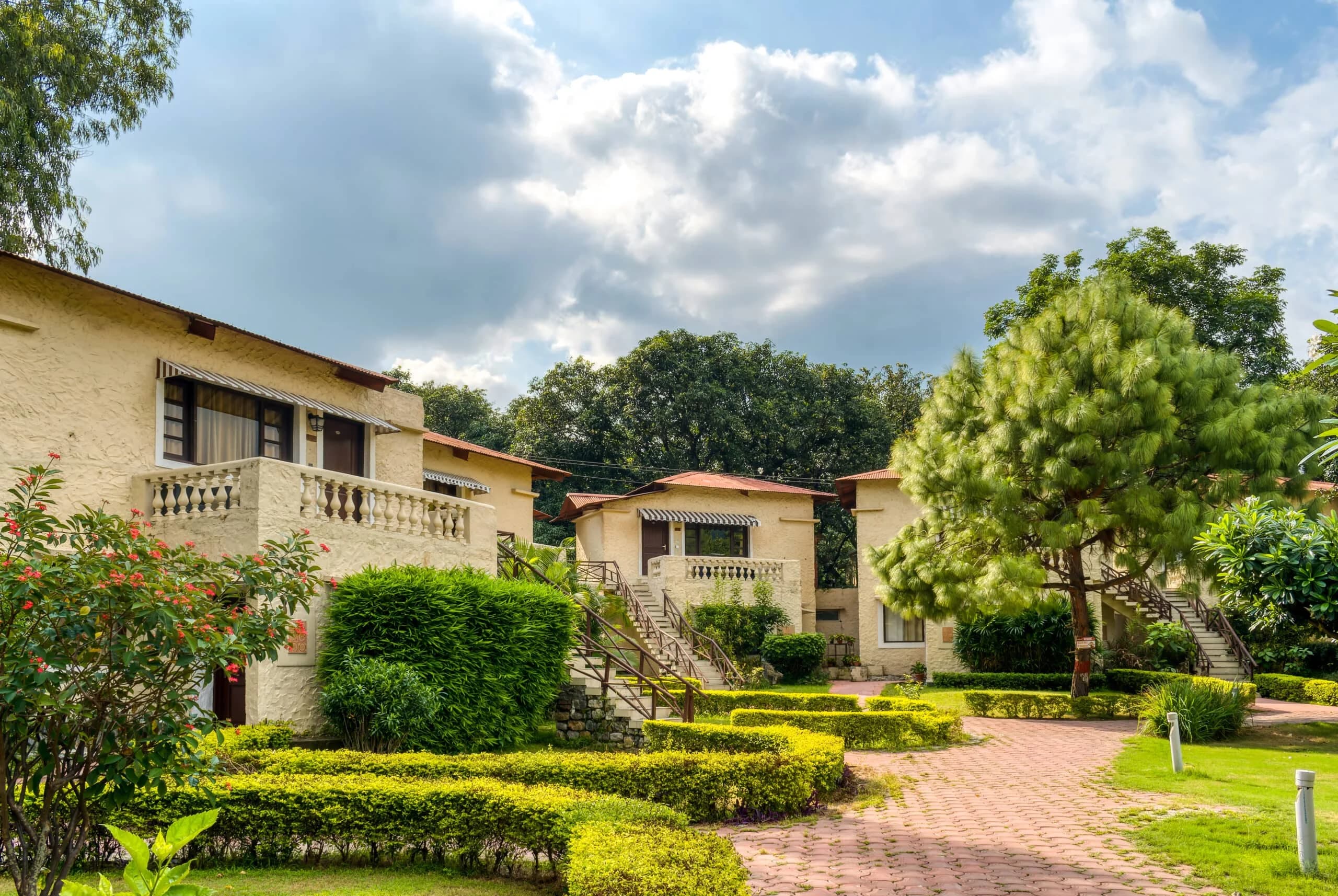 Exterior of resort cottages with pale yellow walls, private balconies, and wooden staircases, surrounded by a lush green lawn and manicured hedges.